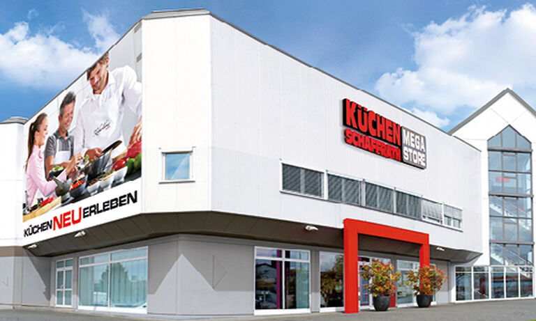 Das Bild zeigt den „Küchen Schaffrath Mega Store“ mit einer weißen Fassade und einem modernen Design. Auf der linken Seite ist ein großes Werbebanner mit einer Gruppe von Menschen zu sehen, die gemeinsam kochen. Darunter steht der Schriftzug „KÜCHEN NEU ERLEBEN“ in schwarzen und roten Buchstaben. Auf der rechten Seite der Fassade prangt das Logo „KÜCHEN SCHAFFRATH MEGA STORE“ in auffälligen roten und weißen Buchstaben. Der Eingangsbereich wird durch einen roten, rechteckigen Rahmen betont, der von zwei Pflanzen in Kübeln flankiert wird. Im Hintergrund ist ein Teil der Fassade aus Glas erkennbar. Der Himmel ist klar und blau, was eine freundliche Atmosphäre vermittelt.